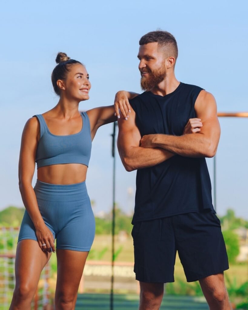 A woman in a blue sports bra and shorts poses next to a man in a black tank top and shorts, both smiling in a sunny outdoor setting.
