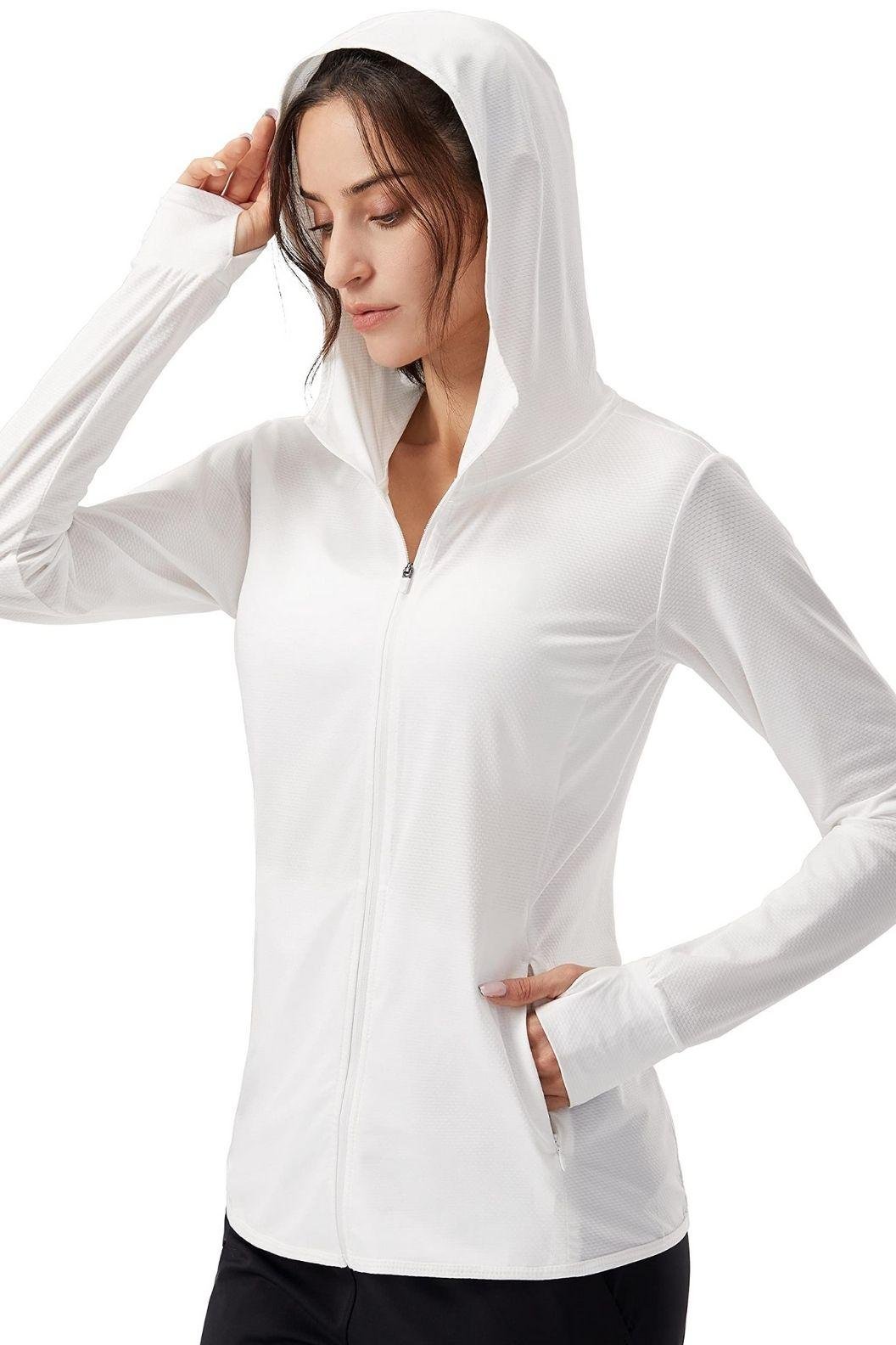 A woman wearing a white zip-up hoodie with a hood, adjusting it while standing in a relaxed pose.